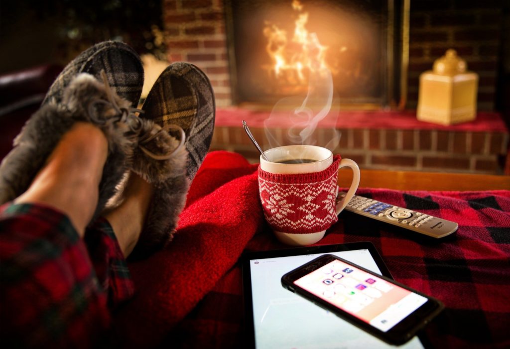 Personne qui se détend au coin du feu les pieds posés sur une table basse sur laquelle il y a une tasse qui fume et un téléphone portable