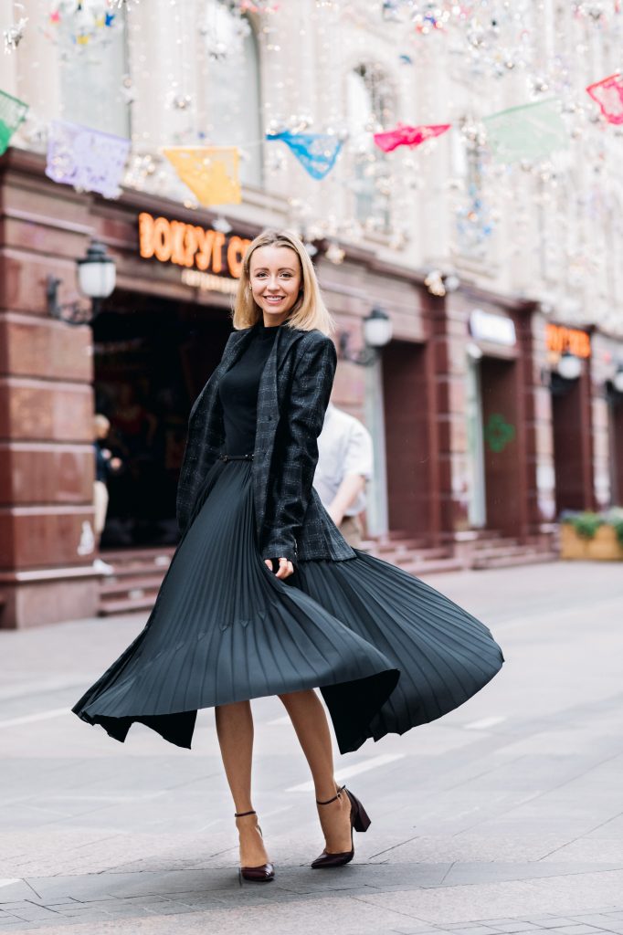 femme blonde dans la rue portant une jupe midi foncée un pull noir et une veste de blazer à carreaux