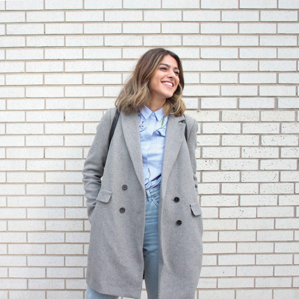 femme portant un manteau long gris un chemisier bleu et un jean devant un mur en briques blanches